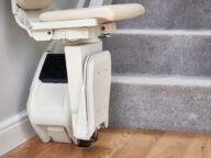 A stairlift is mounted at the base of a carpeted staircase. The stairlift seat is positioned at the bottom, adjacent to wooden flooring.