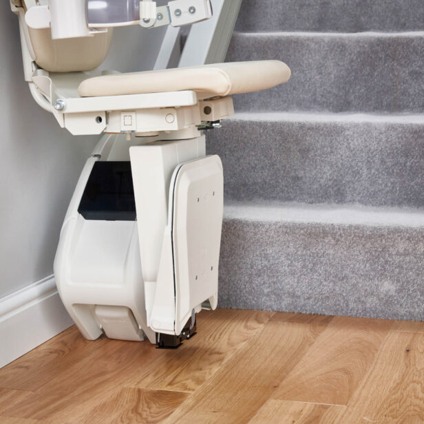 A stairlift is mounted at the base of a carpeted staircase. The stairlift seat is positioned at the bottom, adjacent to wooden flooring.