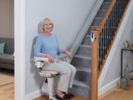 A woman sits on a stairlift at the bottom of a staircase in a well-lit living room.