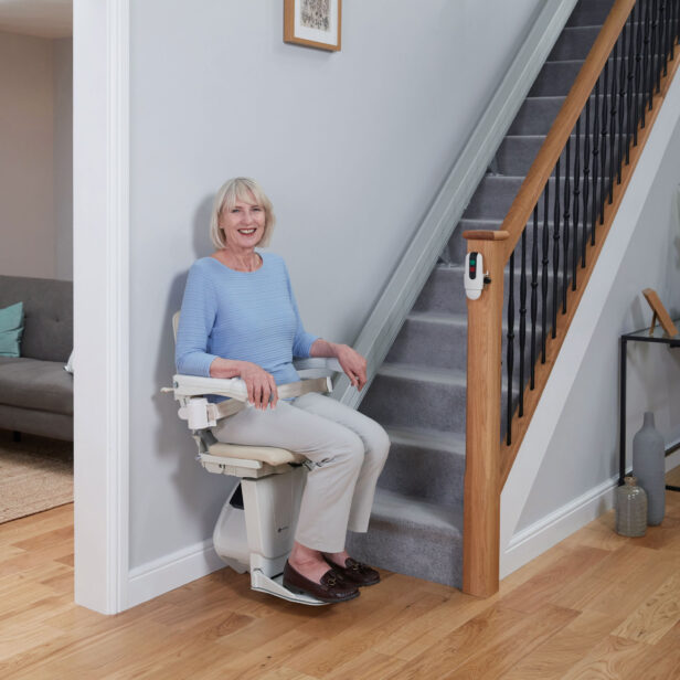 A woman sits on a stairlift at the bottom of a staircase in a well-lit living room.