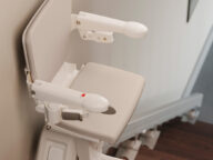 A beige stairlift is attached to a staircase, featuring a padded seat, armrests, and a safety belt.