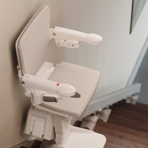 A beige stairlift is attached to a staircase, featuring a padded seat, armrests, and a safety belt.