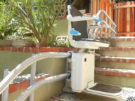 Outdoor stair lift with a cushioned seat and armrests installed on brick steps, designed to assist individuals in navigating stairs. Brushes and natural oak wood fence are visible in the background.