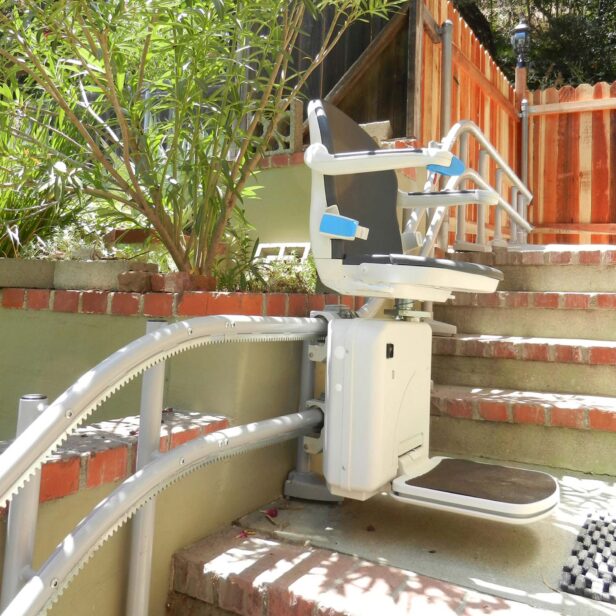 Outdoor stair lift with a cushioned seat and armrests installed on brick steps, designed to assist individuals in navigating stairs. Brushes and natural oak wood fence are visible in the background.