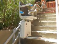 A stair lift is installed along an outdoor flight of stairs, featuring a chair with armrests and a footrest on a rail system for accessibility.