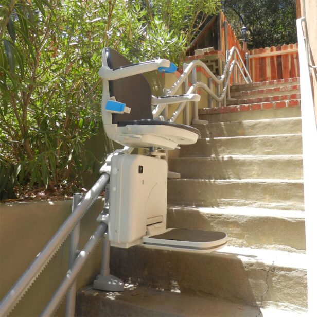 A stair lift is installed along an outdoor flight of stairs, featuring a chair with armrests and a footrest on a rail system for accessibility.