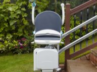 Outdoor stair lift installed beside a wooden staircase in a garden, with greenery and flowers in the background.