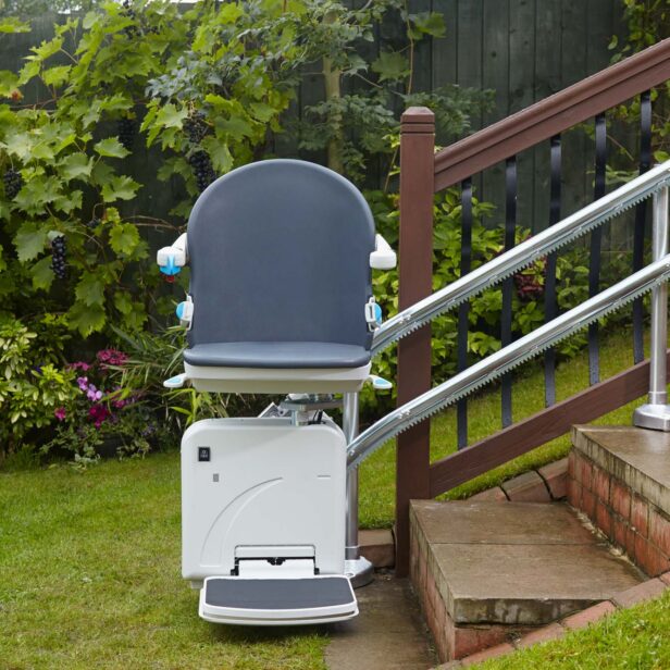 A stair lift is installed along an outdoor staircase, bordered by greenery and a wooden railing.