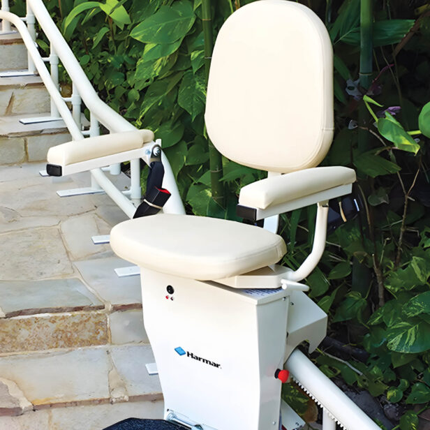 A stairlift installed beside an outdoor stone stairway, designed to assist with mobility. The stairlift has a cushioned seat, armrests, and is positioned next to greenery.