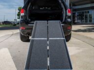 A black SUV with its rear hatch open and two portable aluminum ramps extending from the cargo area to the ground, positioned on a paved surface.