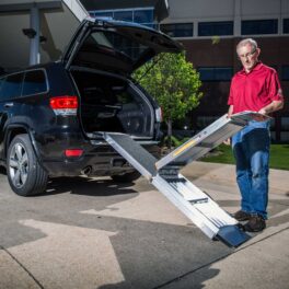 A man stands behind an SUV, holding a partially unfolded aluminum ramp extending from the vehicle's open trunk.