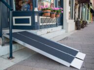 A portable ramp provides wheelchair access to a storefront with steps. The ramp leads from the sidewalk to the entrance, which is decorated with potted flowers.