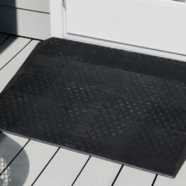 A black rubber doormat with a textured pattern is placed in front of a white door on a light gray wooden deck.