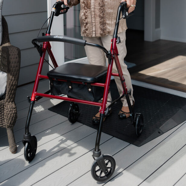 A person using a red walker is entering a house via a ramp from a wooden deck. The walker has a seat and four wheels.