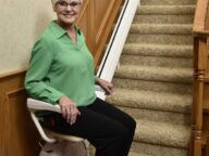 A woman with short white hair and glasses sits on a motorized stairlift at the base of a carpeted staircase. She is dressed in a green blouse and black pants, smiling at the camera.