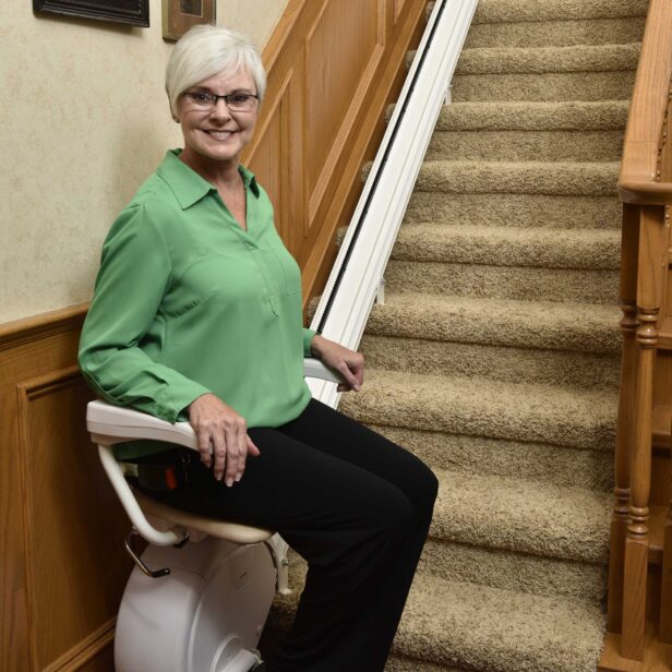 A woman with short white hair and glasses sits on a motorized stairlift at the base of a carpeted staircase. She is dressed in a green blouse and black pants, smiling at the camera.