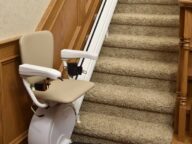 A stair lift is installed on a carpeted staircase with wooden railings, featuring a padded seat, armrests, and a footrest.