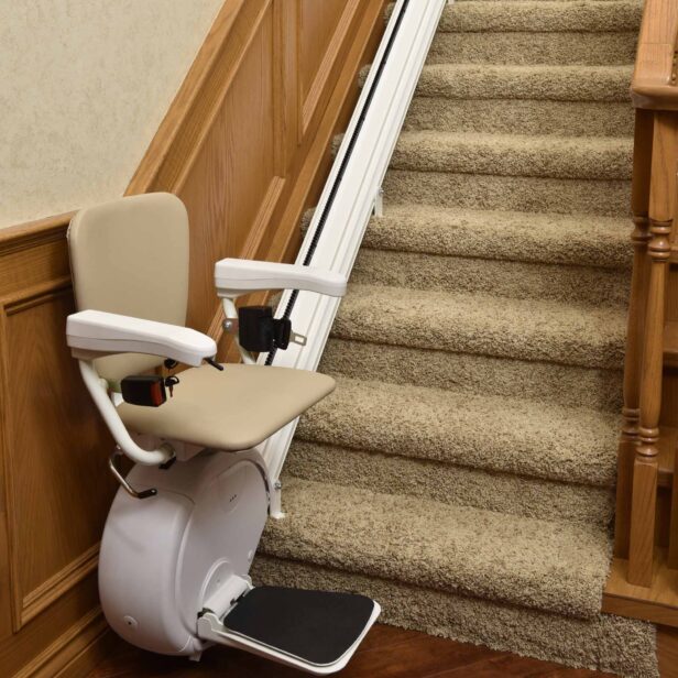 A stair lift is installed on a carpeted staircase with wooden railings, featuring a padded seat, armrests, and a footrest.