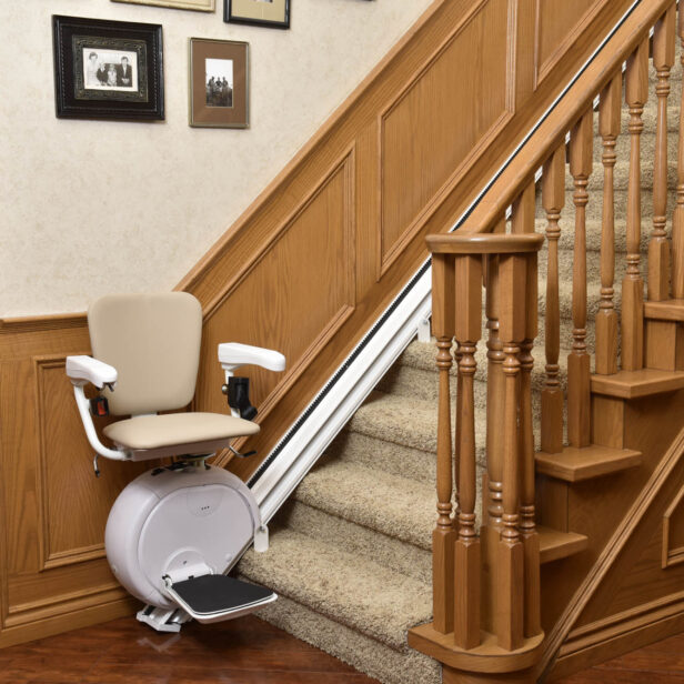 A stairlift installed along a carpeted staircase with wooden railings in a home, featuring a beige seat and a footrest.