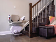 A white and gray stair lift is installed along a wooden staircase next to a gray wall. An armchair is partially visible next to the staircase.