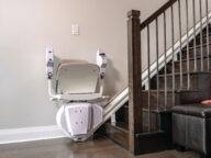 A stair lift chair is mounted on a rail along a wooden staircase, positioned at the bottom of the steps, next to a wall and a black leather ottoman.