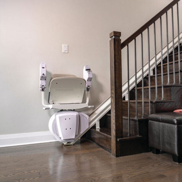 A stair lift chair is mounted on a rail along a wooden staircase, positioned at the bottom of the steps, next to a wall and a black leather ottoman.