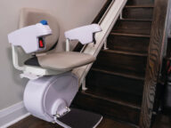 A stair lift is installed alongside a wooden staircase, providing a seat with armrests and footrest on a rail system for assisting with mobility.