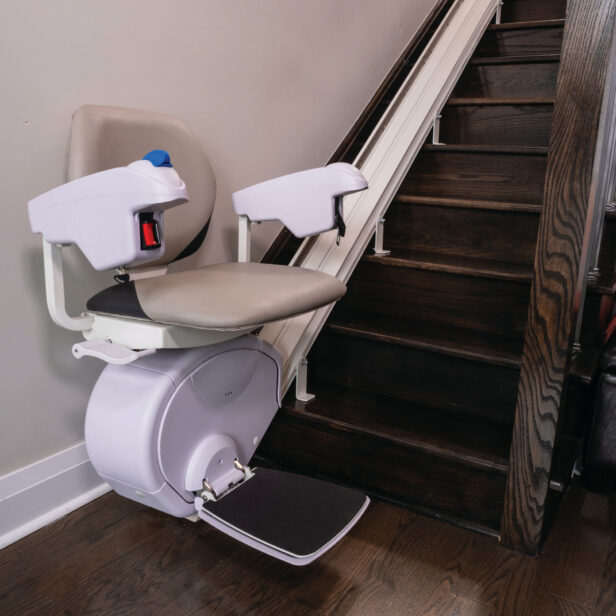 A stair lift is installed alongside a wooden staircase, providing a seat with armrests and footrest on a rail system for assisting with mobility.