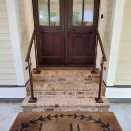 A brick staircase with metal handrails leads to a dark wooden double door. A doormat with a partial design sits at the base of the steps.