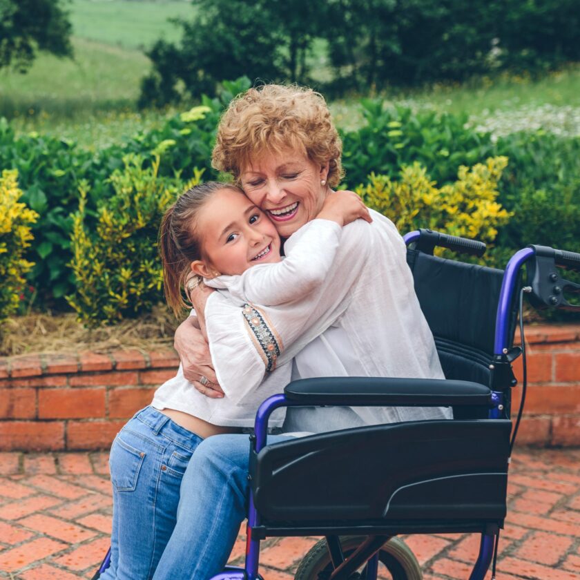 A girl hugs an elderly woman sitting in a wheelchair. They are outdoors with greenery and a brick walkway in the background.