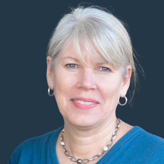 A person with short grey hair and hoop earrings is wearing a blue top and a necklace, posing against a dark background.