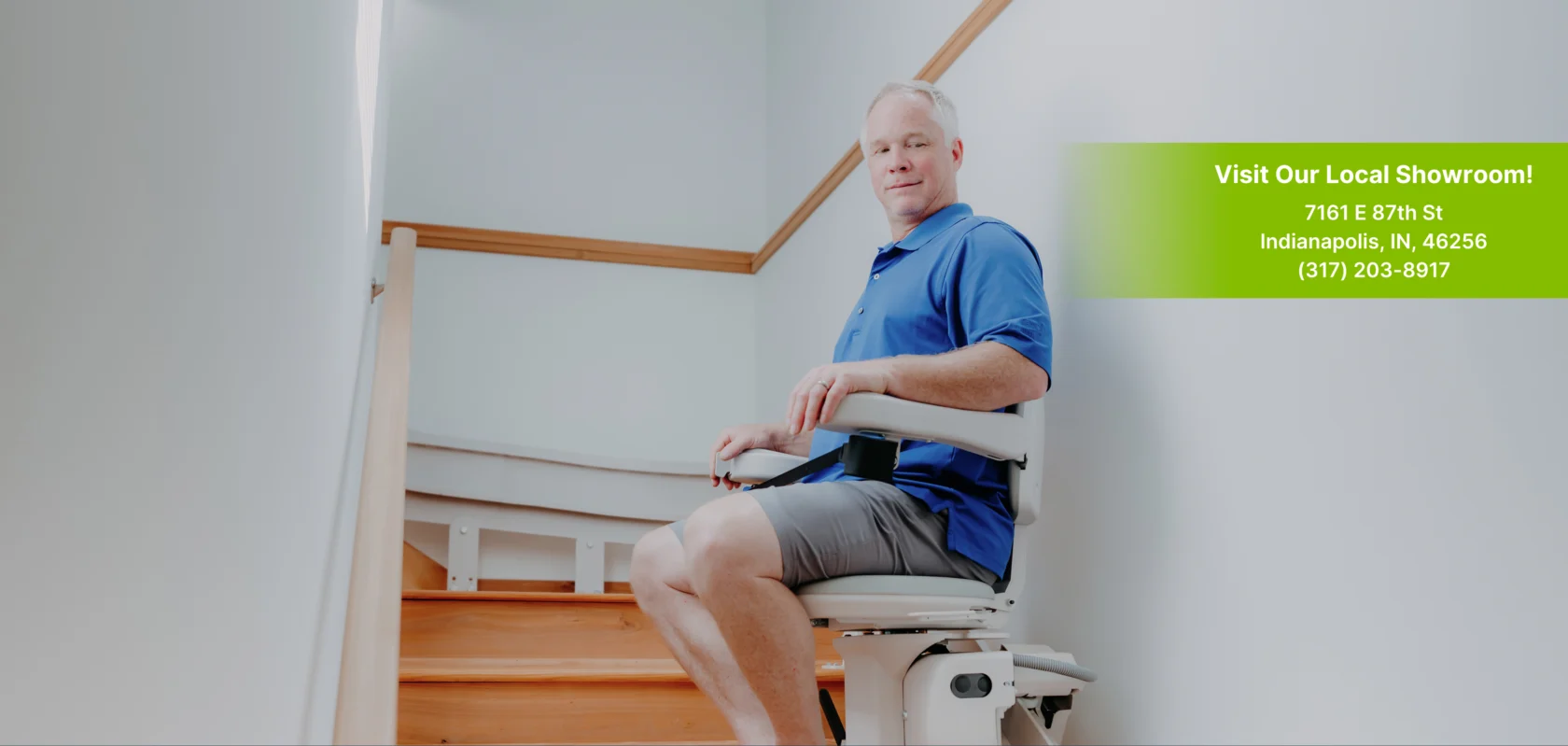 A man in a blue shirt sits on a stair lift, with a showroom address and phone number displayed on a green overlay to his right.