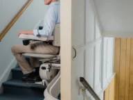 A man sits on a stair lift traveling up a staircase in a home. The stair lift is attached to a rail along the wall.