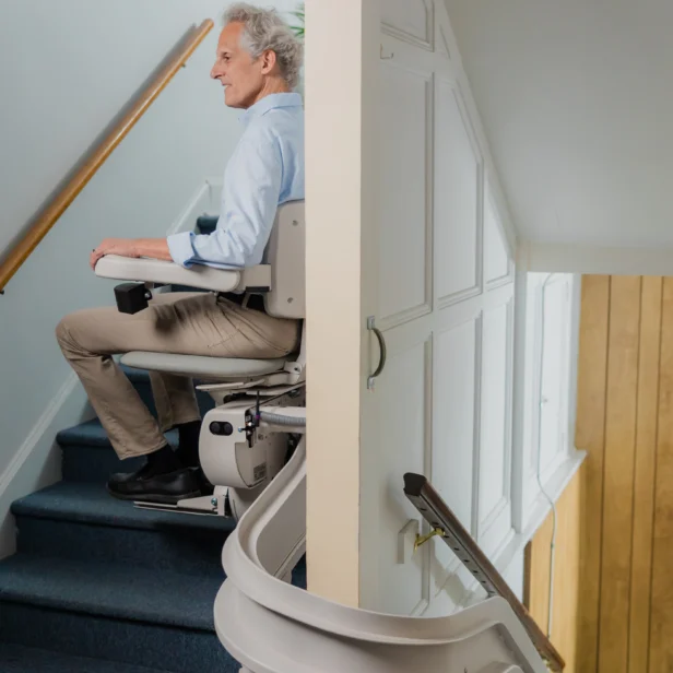 A man sits on a stair lift traveling up a staircase in a home. The stair lift is attached to a rail along the wall.