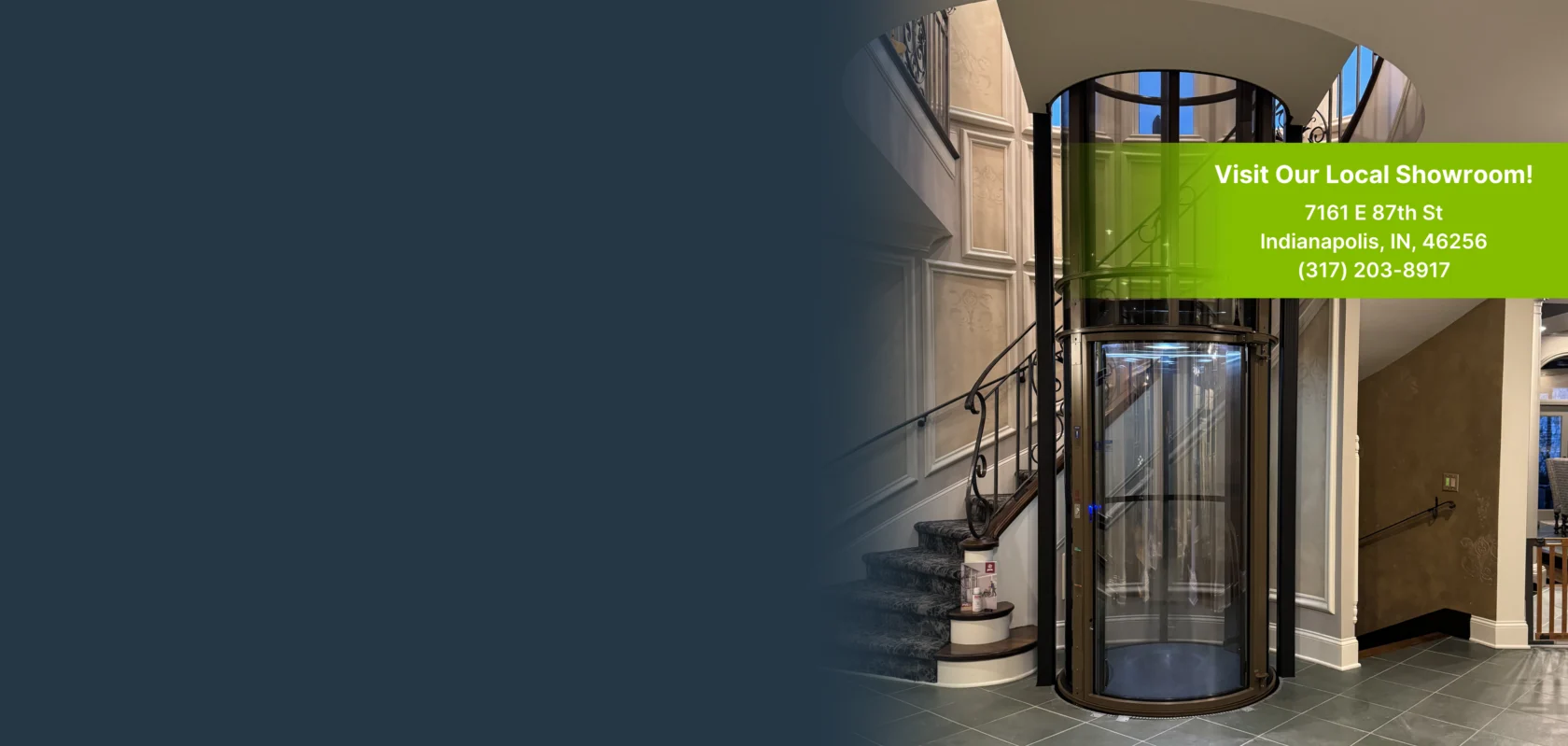 Glass elevator in a showroom with a staircase. A green sign displays showroom address: 7161 E 87th St, Indianapolis, IN, 46256, and phone number: (317) 203-8917.