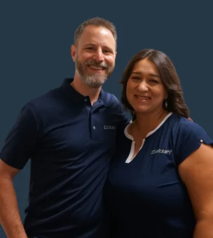 A man and woman wearing dark blue shirts with company logos stand together, both smiling, against a solid dark background.