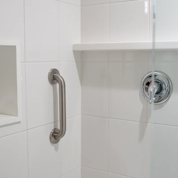 A bathroom shower with white tile walls, a stainless steel grab bar, and a single-lever handle for the shower controls.