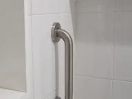 A stainless steel grab bar is mounted vertically on a white tiled bathroom wall near a recess shelf.