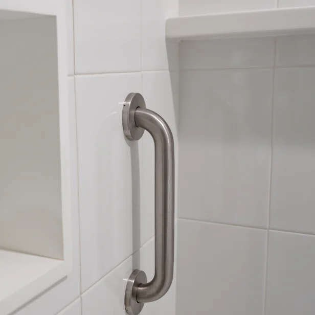 A stainless steel grab bar is mounted vertically on a white tiled bathroom wall near a recess shelf.