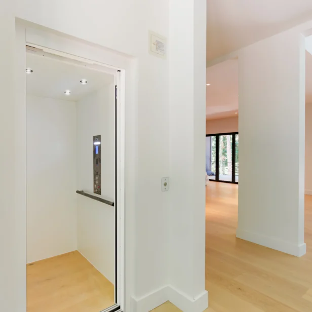 A modern residential elevator with open doors, located in a bright, spacious interior with wooden flooring and white walls.