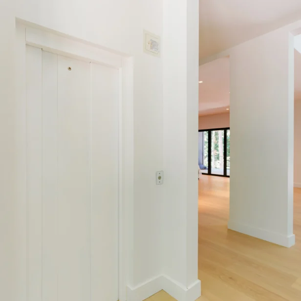 A white hallway with light wooden flooring features a closed elevator door on the left and large windows in the background.