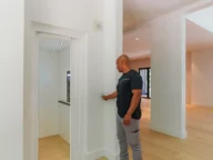 A man stands in a modern, minimally furnished room, pressing a button next to an open elevator door.