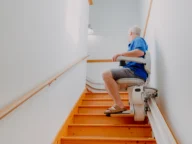 A person in a blue shirt and shorts rides a stairlift up a wooden staircase in a well-lit home.