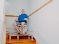 A man sits on a stairlift, moving up a wooden staircase in a brightly lit indoor setting.