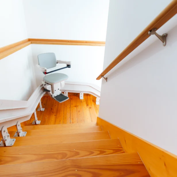 A stairlift installed along a wooden staircase with white walls and wooden handrails, designed for assisting individuals with mobility issues in navigating the stairs.