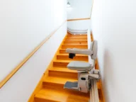 A stairlift is installed on a wooden staircase in a brightly lit, narrow stairway with white walls and wooden handrails.