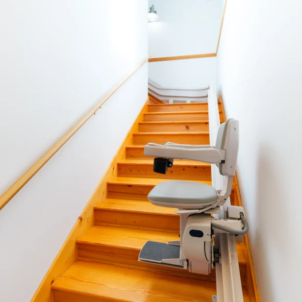 A stairlift is installed on a wooden staircase in a brightly lit, narrow stairway with white walls and wooden handrails.