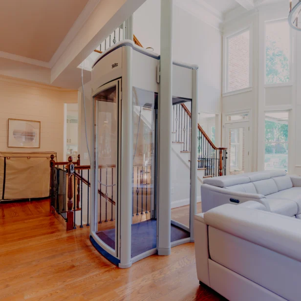 A small glass elevator is situated in the center of a spacious, brightly lit living room with a large gray sectional sofa, wooden floors, and a staircase leading to an upper level.