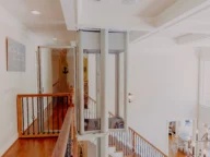 A modern home interior with wooden floors and an indoor elevator connecting the upper and lower levels, surrounded by metal railings and well-lit by natural light.