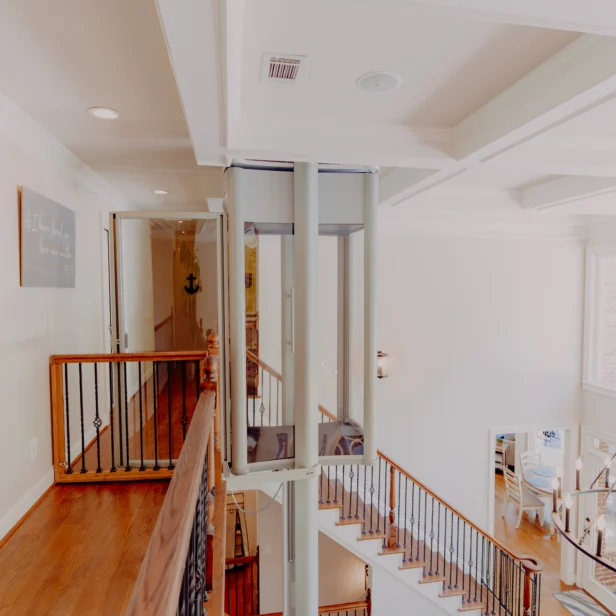 A modern home interior with wooden floors and an indoor elevator connecting the upper and lower levels, surrounded by metal railings and well-lit by natural light.